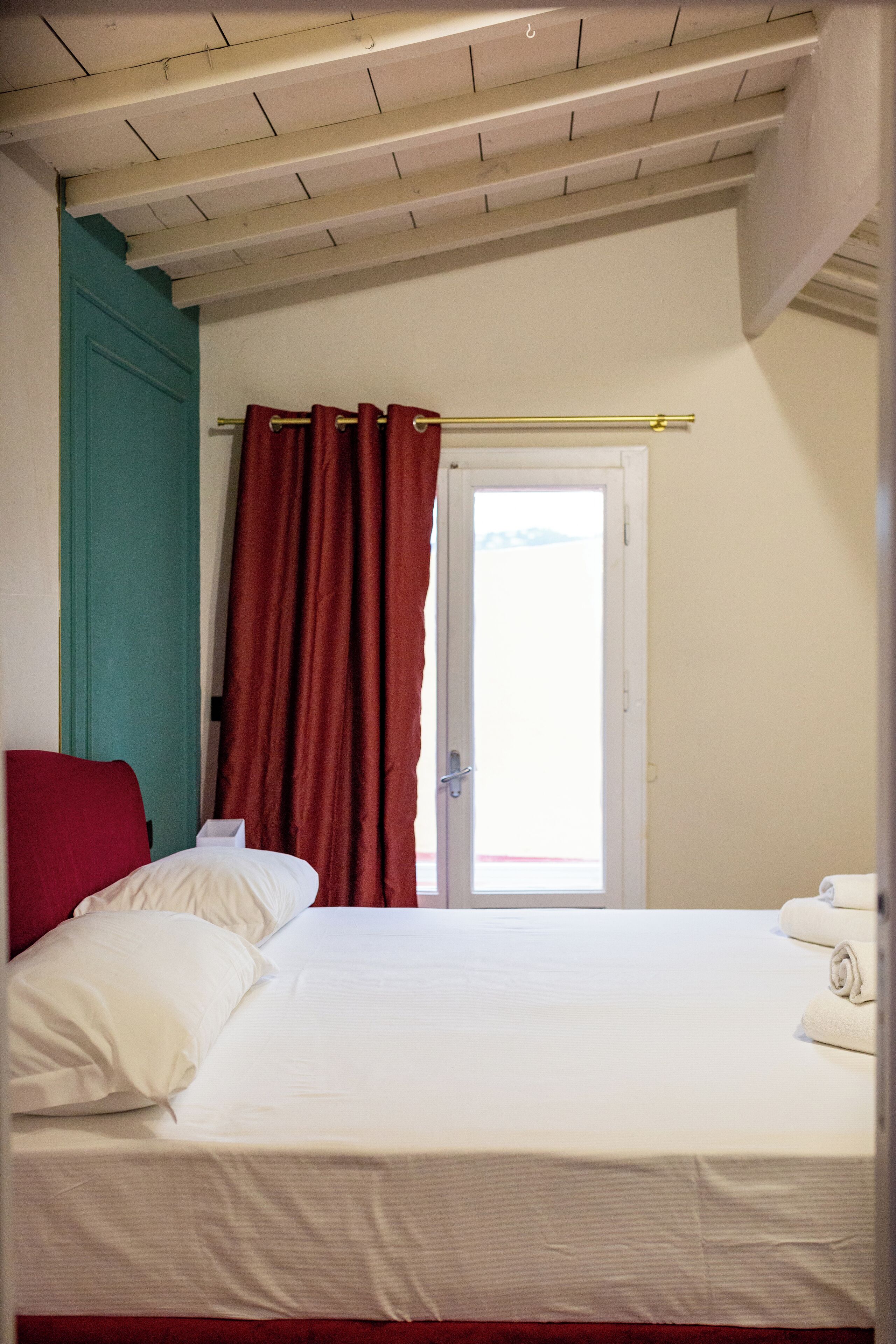 Habitació d'hotel amb un llit, una cadira vermella, una porta verda blavosa i una finestra amb cortines vermelles.