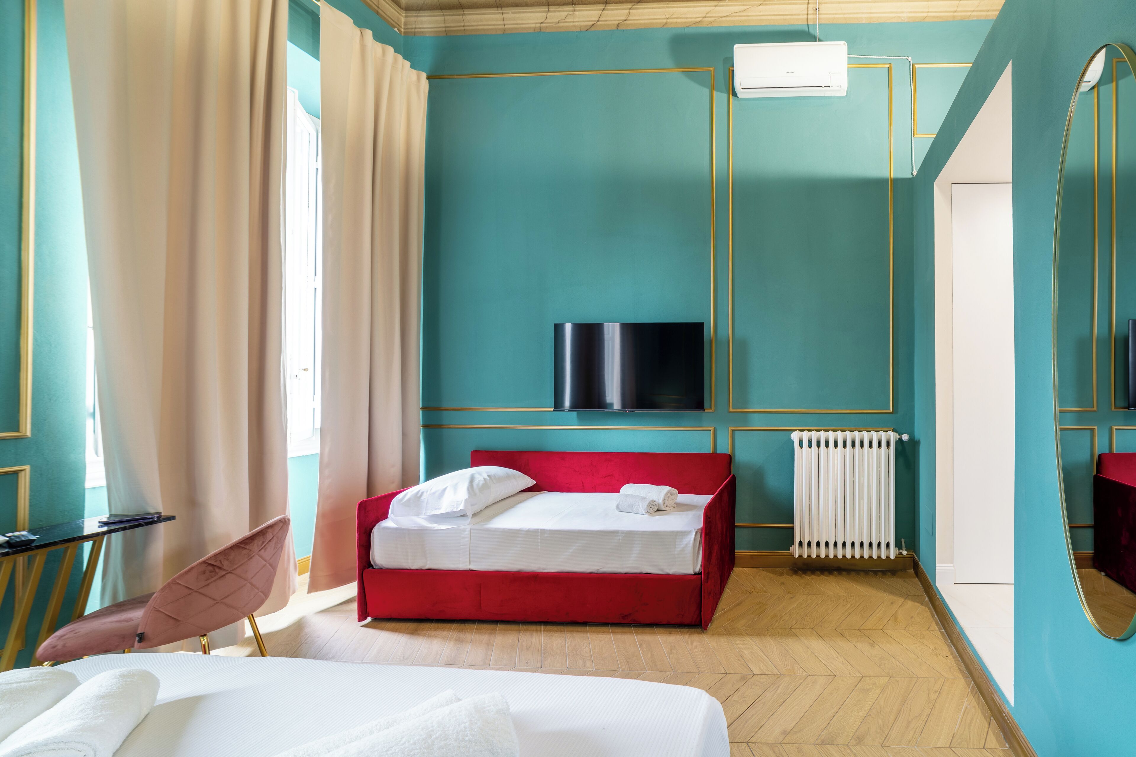 A hotel room with a red bed, a TV mounted on the wall, a white radiator, and a large mirror.