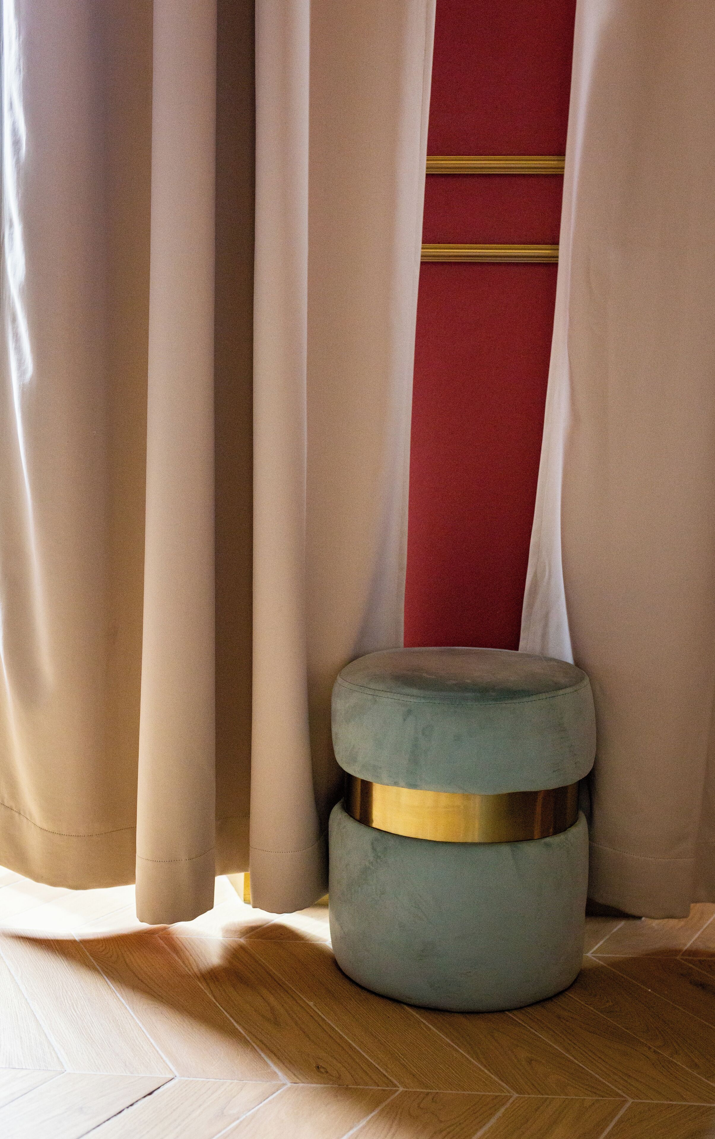 Une pièce avec un plancher en bois, un pouf gris orné d’une bande dorée et un rideau rouge et beige.