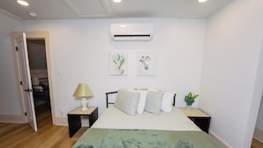 Une chambre à coucher comprenant un lit, des tables de chevet, une lampe, un climatiseur et deux tableaux accrochés au mur.