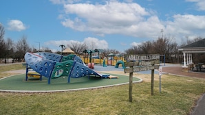 Une aire de jeux avec un toboggan et des structures d’escalade, entourée d’un parc agrémenté de bancs et d’un kiosque.