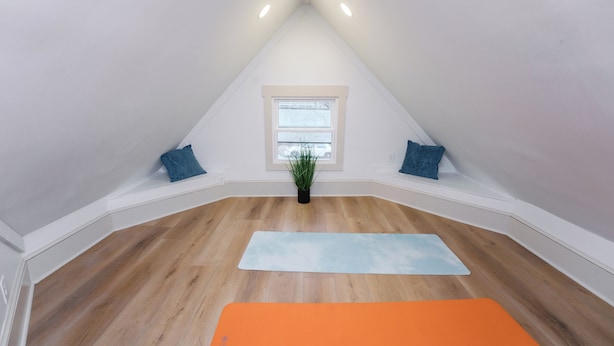Une pièce avec un plafond en pente, une fenêtre avec des stores, une plante en pot et deux tapis de yoga.