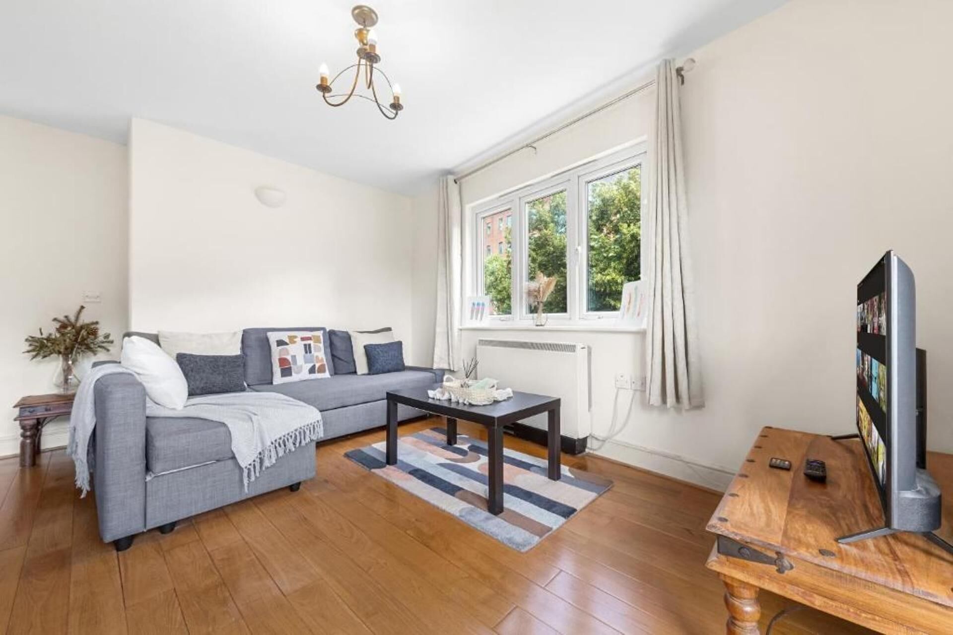 A living room with a grey sofa, a wooden coffee table, a TV on a wooden stand, and a window with curtains.