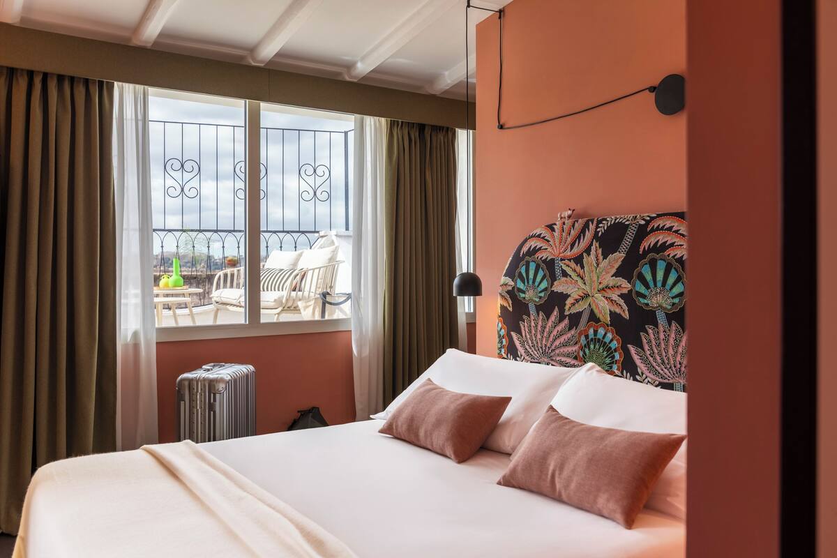 A hotel room with a bed, curtains, a window with a balcony view, and a patterned headboard.
