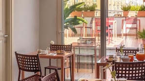 Un espace repas avec des tables et des chaises, une fenêtre donnant sur des bâtiments et des plantes, et une porte située à gauche.