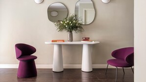Une pièce avec une table blanche, deux chaises violettes, un vase avec des fleurs et deux miroirs fixés au mur.