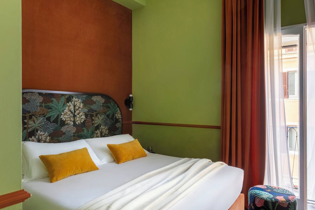 A bed with white bedding and two yellow pillows, a headboard with a floral pattern, and a view of a building through a window with red curtains.