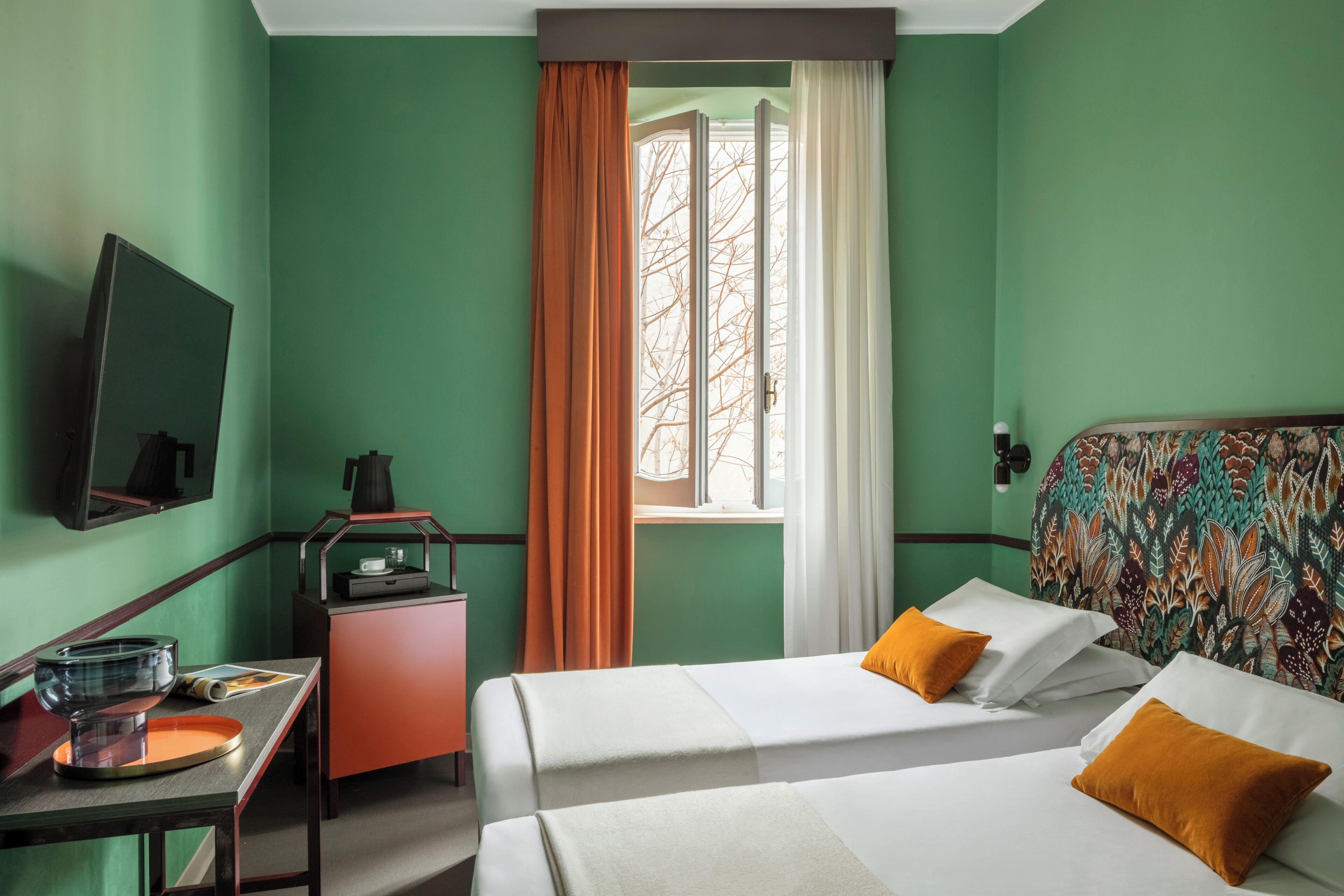 Ein Hotelzimmer mit zwei Betten, einem Schreibtisch mit Wasserkocher, einem Spiegel und einem Fenster mit Vorhängen.