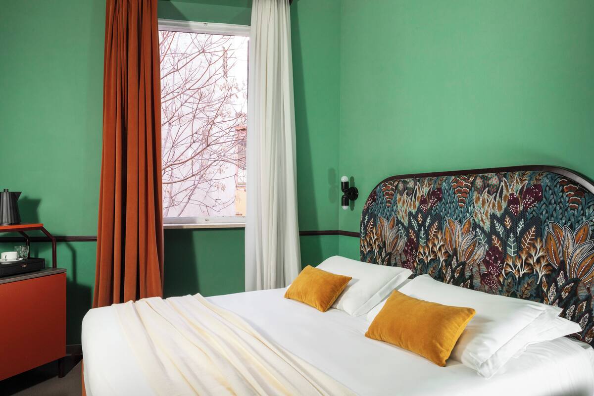 A hotel room with a bed, a nightstand, a window with curtains, and a patterned headboard.