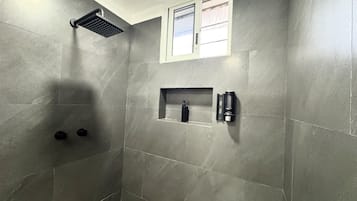 Un área de ducha con cabezal de ducha tipo lluvia empotrado, una ventana pequeña y un estante con artículos de aseo.