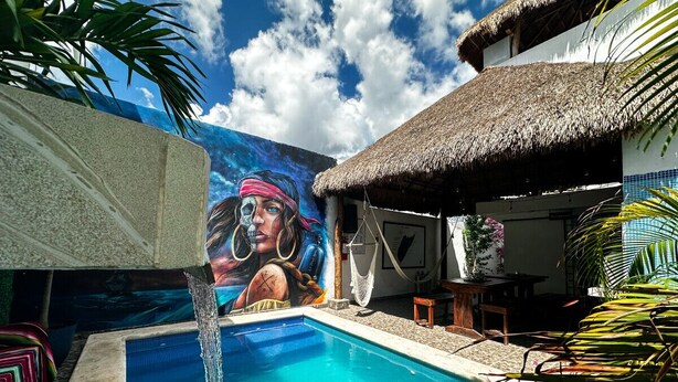 Una piscina con un mural, una estructura de techo de paja y plantas tropicales.