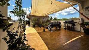 Un patio amplio con suelo de madera, una pérgola colgante, mobiliario de exterior y plantas en macetas.