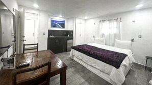 Una habitación con cama, escritorio, silla, televisión y kitchenette.