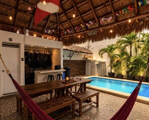 Zona junto a la piscina con mesa y bancos de madera, techo de paja y una piscina.