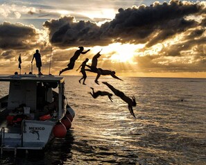 A boat with the word 'MEXICO' on the side, people jumping into the water, and a sunset in the background. - La Perle Noire | HM - Terasse et Piscine (San Miguel de Cozumel)