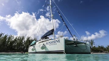 A sailboat named 'SUNSHINE' with a registration number 'MAT DL2362AP' anchored near a beach with clear turquoise water and a backdrop of trees and a partly cloudy sky.