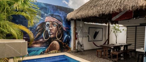 A swimming pool with a mural, a thatched-roof structure, and outdoor seating.