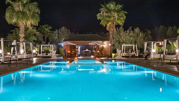 Una piscina notturna con sedie a sdraio e ombrelloni, circondata da palme e un edificio con un portico coperto.