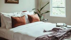 A neatly made bed with white linens and a striped blanket, a framed picture on the wall, a potted plant, and a window with a view of greenery. - Luxury 5BR Townhome w/ Private Plunge Pool in Lower Garden District (New Orleans)