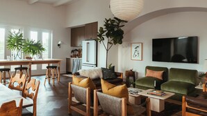 A spacious living room with a large TV, comfortable seating, a dining area with wooden chairs, and a kitchen in the background. - Luxury 5BR Townhome w/ Private Plunge Pool in Lower Garden District (New Orleans)