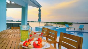 A wooden table with a plate of skewered vegetables and a pitcher of iced tea, overlooking a swimming pool and a body of water during sunset. - New Waterfront house pool hot tub golf cart kayaks (Indian Rocks Beach)
