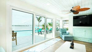 A spacious living area with large sliding glass doors leading to a poolside area, a ceiling fan, and a television. - New Waterfront house pool hot tub golf cart kayaks (Indian Rocks Beach)
