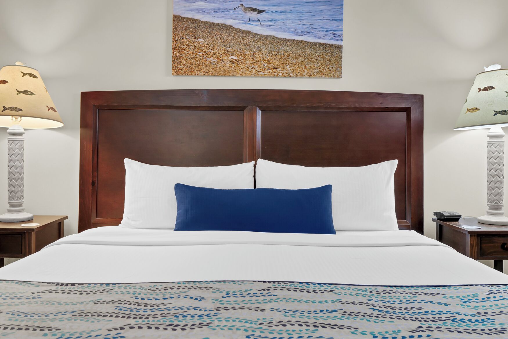 A neatly made bed with white and blue bedding, a wooden headboard, and a beach-themed painting above it.