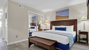 A hotel room with a large bed, a bedside table, a nightstand, a mirror, and a view of the beach.