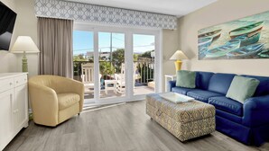 Un salon moderne avec un fauteuil beige, un canapé bleu et un pouf à motifs. Un tableau représentant des bateaux est accroché au mur, et un balcon offre une vue sur la mer.