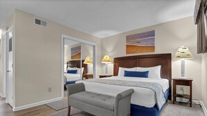 A hotel room with a large bed, two bedside lamps, a bench, a painting on the wall, and a door leading to another room. - Summer Sands Diggin' In The Sand Room 103 (Wrightsville Beach)