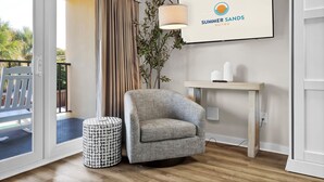 Ein Zimmer mit grauem Sessel, rundem Hocker, einer Lampe und einem gerahmten Schild mit der Aufschrift 'Summer Sands Suites'.