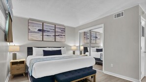 A modern bedroom with a large bed, bedside tables, a bench, and artwork on the walls. - Summer Sands- Isle Be Back Room 203 (Wrightsville Beach)