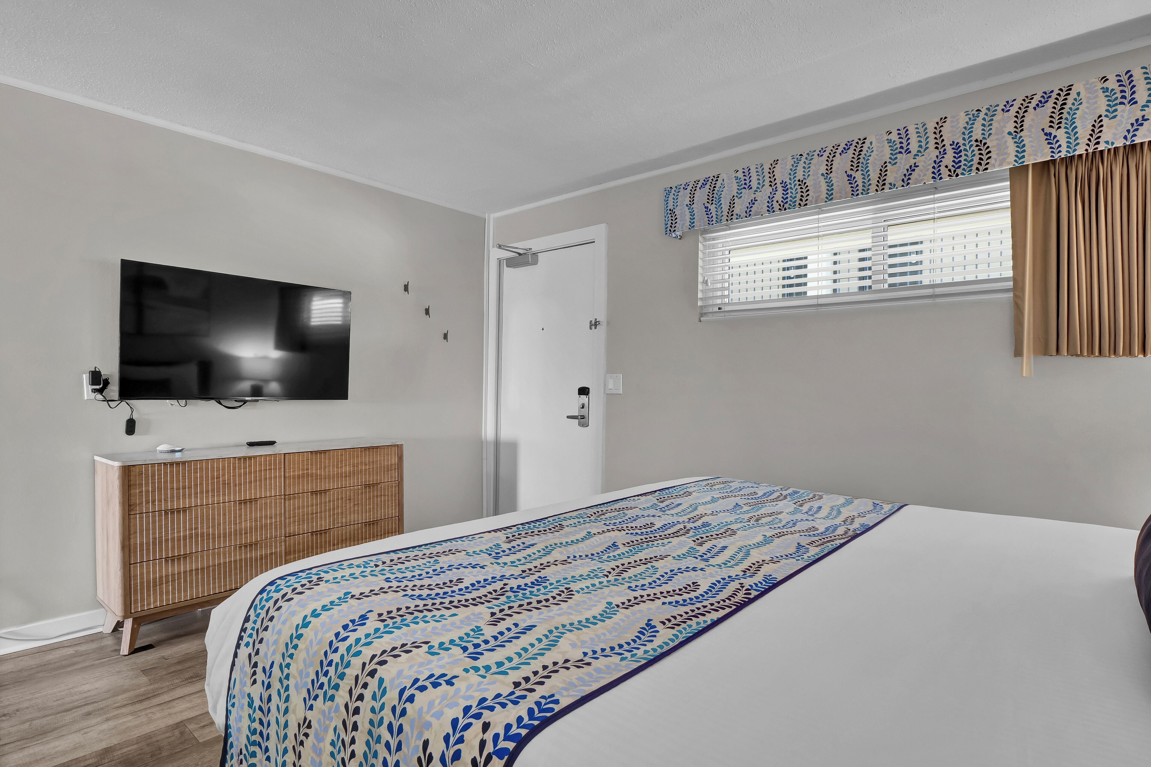 Une chambre à coucher moderne avec un grand lit, un téléviseur fixé au mur, une armoire en bois et une fenêtre avec des stores.