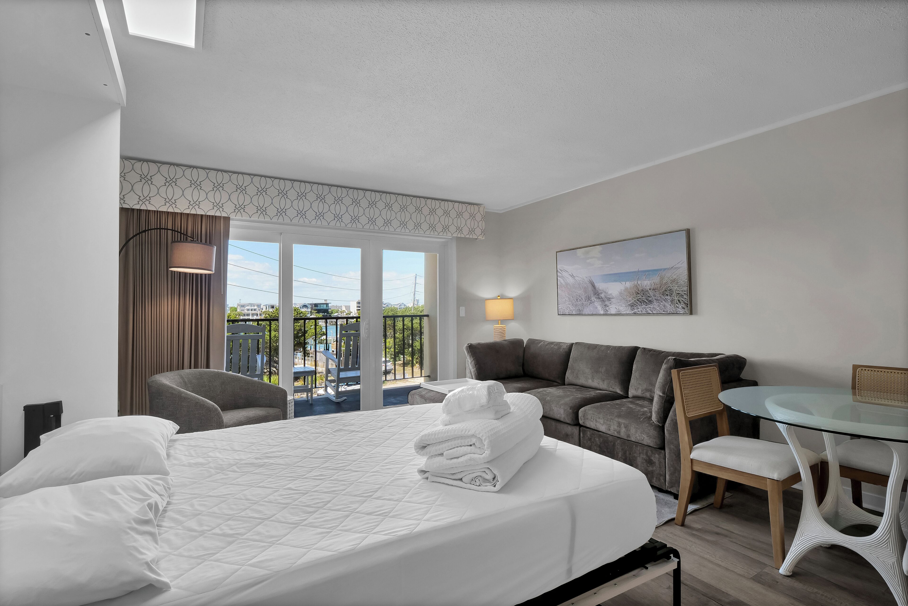 A hotel room with a bed, a sofa, a dining table with chairs, a balcony with a view, and a painting on the wall.