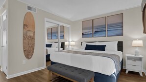 Quarto com uma cama grande, um banquinho, uma prancha de surf de madeira, um espelho e vista para o oceano.