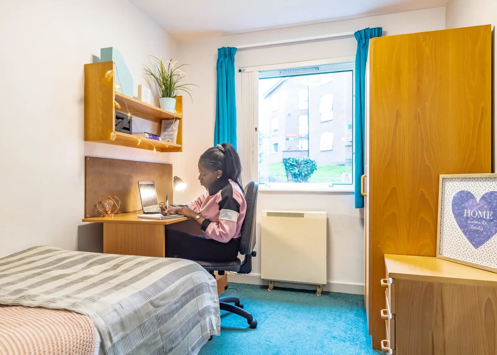 A room with a bed, a desk, a chair, a radiator, a window with curtains, and a shelf with a plant.