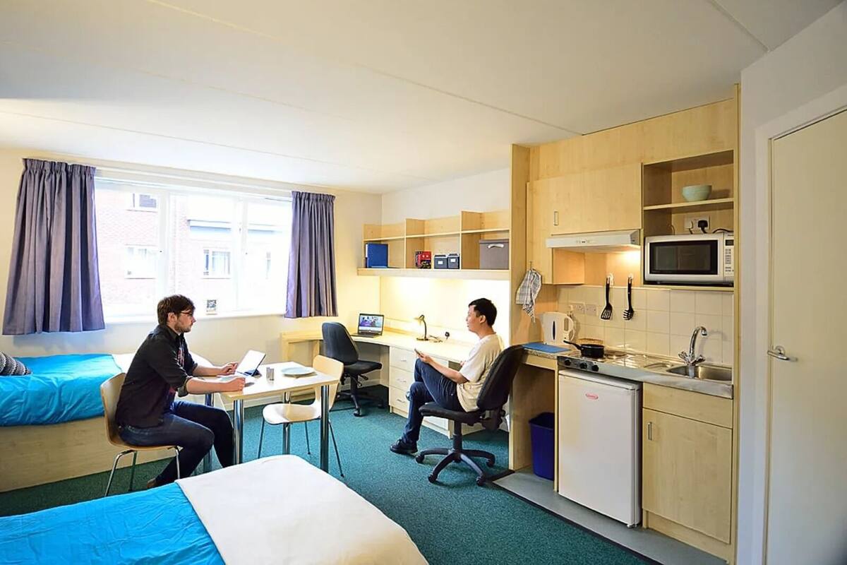Habitación de hotel compacta con dos camas, kitchenette y escritorio con computadoras.