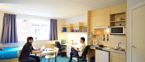 Habitación de hotel compacta con dos camas, kitchenette y escritorio con computadoras.