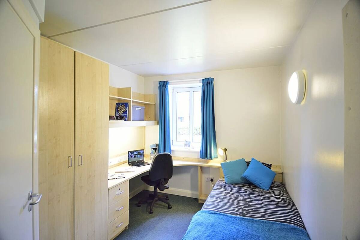 Habitación tipo dormitorio con una cama individual, un escritorio con silla, un armario y una ventana con cortinas.