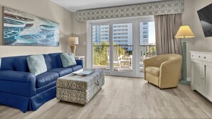 A modern living room with a blue sofa, beige armchair, and patterned ottoman. A painting of boats is on the wall, and there is a balcony with a view of buildings and greenery. - Summer Sands- Lazy Dog Days (Wrightsville Beach)