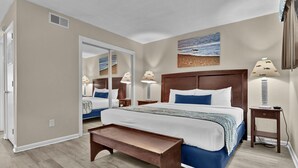 A bedroom with a large bed, two bedside tables, a bench, a mirror, and a painting on the wall. - Summer Sands- Lazy Dog Days (Wrightsville Beach)