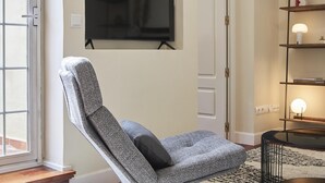 Una sala moderna con un sillón de color gris con estampado, un televisor de pantalla plana montado en la pared, una estantería con objetos decorativos y una lámpara de pie.