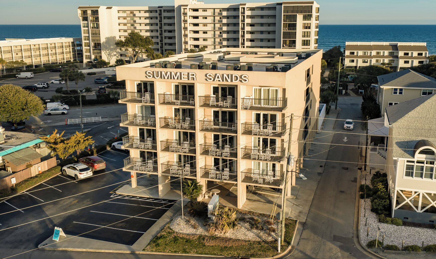 A multi-story hotel named 'Summer Sands' with balconies, a parking lot, and a view of the sea.