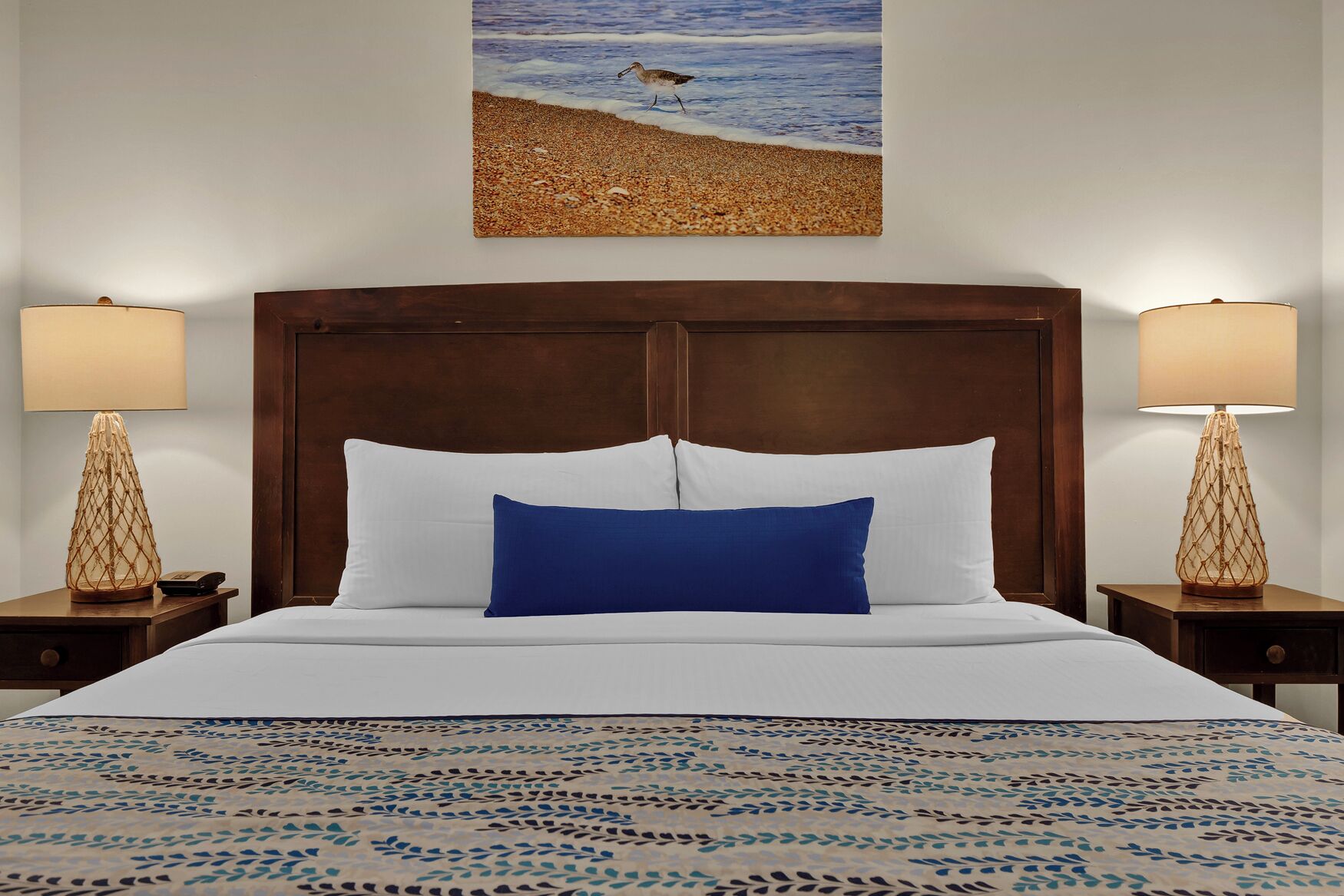 A hotel room with a large bed, two bedside lamps, a wooden headboard, and a beach-themed painting on the wall.