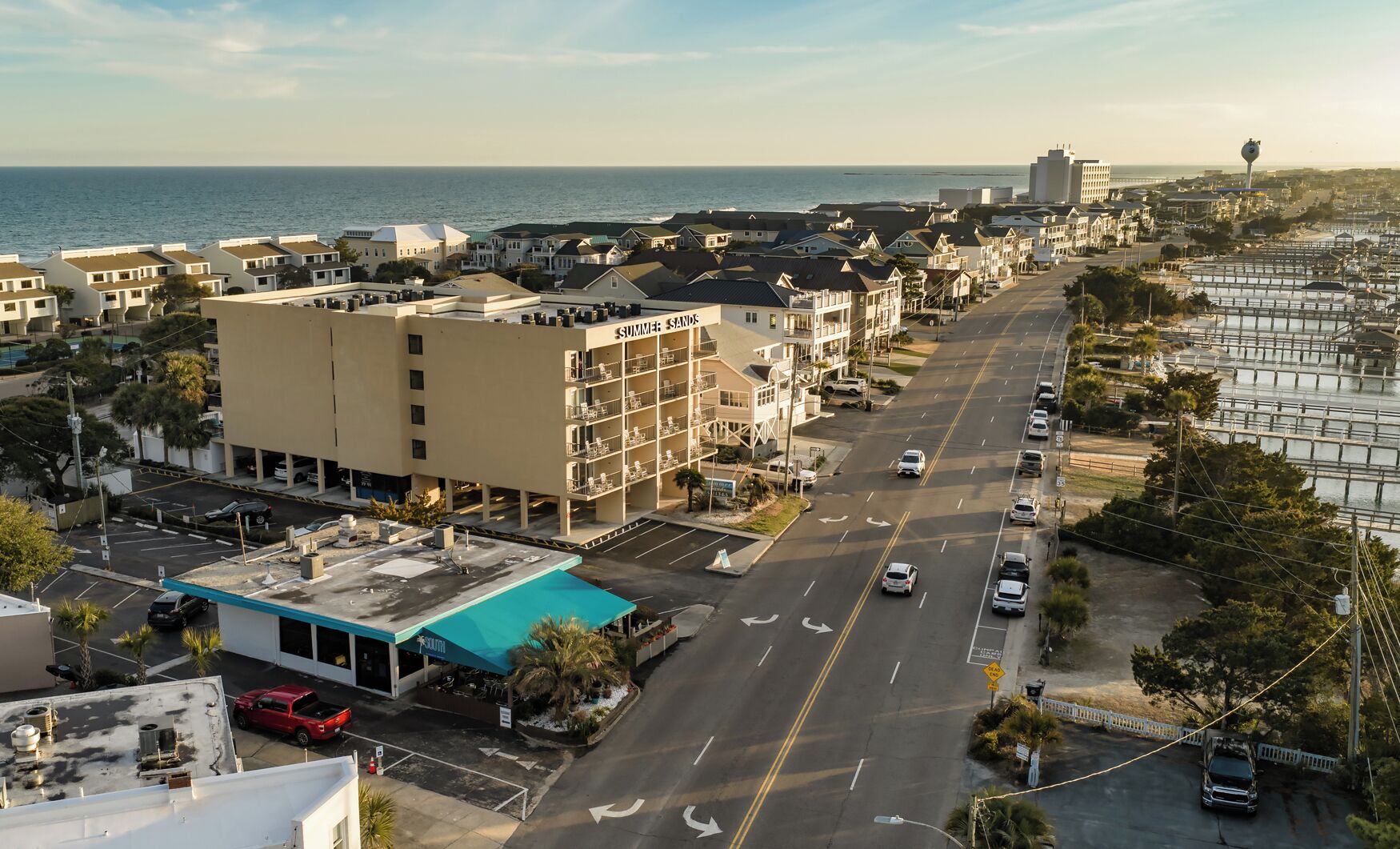 A coastal street with multi-story buildings, a parking lot, and a view of the ocean.