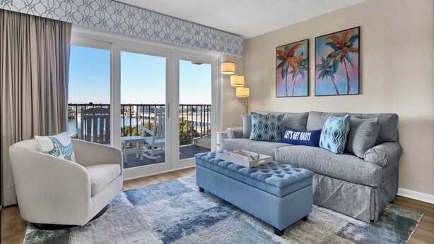 Un salon moderne avec un canapé gris, un fauteuil blanc et un pouf bleu. Deux tableaux ornent le mur, il y a un balcon avec vue sur une piscine, et une grande fenêtre avec des rideaux.