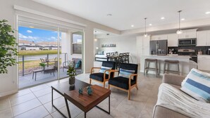 A modern kitchen with white cabinets, a microwave, and a refrigerator. There is a dining area with a table and chairs, and a balcony with a view of the countryside.