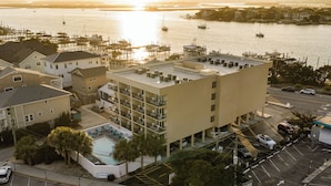 Un hotel de varios pisos con piscina en la azotea, un puerto deportivo al fondo y un estacionamiento con varios automóviles.