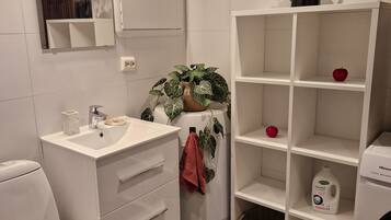 Un baño moderno con un lavamanos blanco, espejo e inodoro. Hay una repisa con una planta en maceta y una manzana roja.