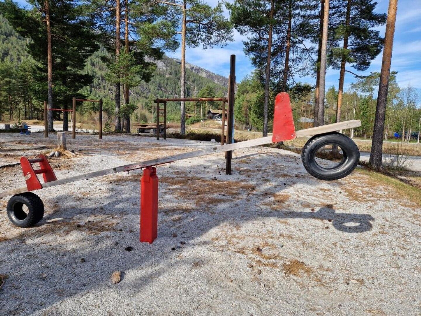 Zona de jocs amb cadira de balanç, suports per penjar i una estructura per pujar, tot en una zona boscosa.
